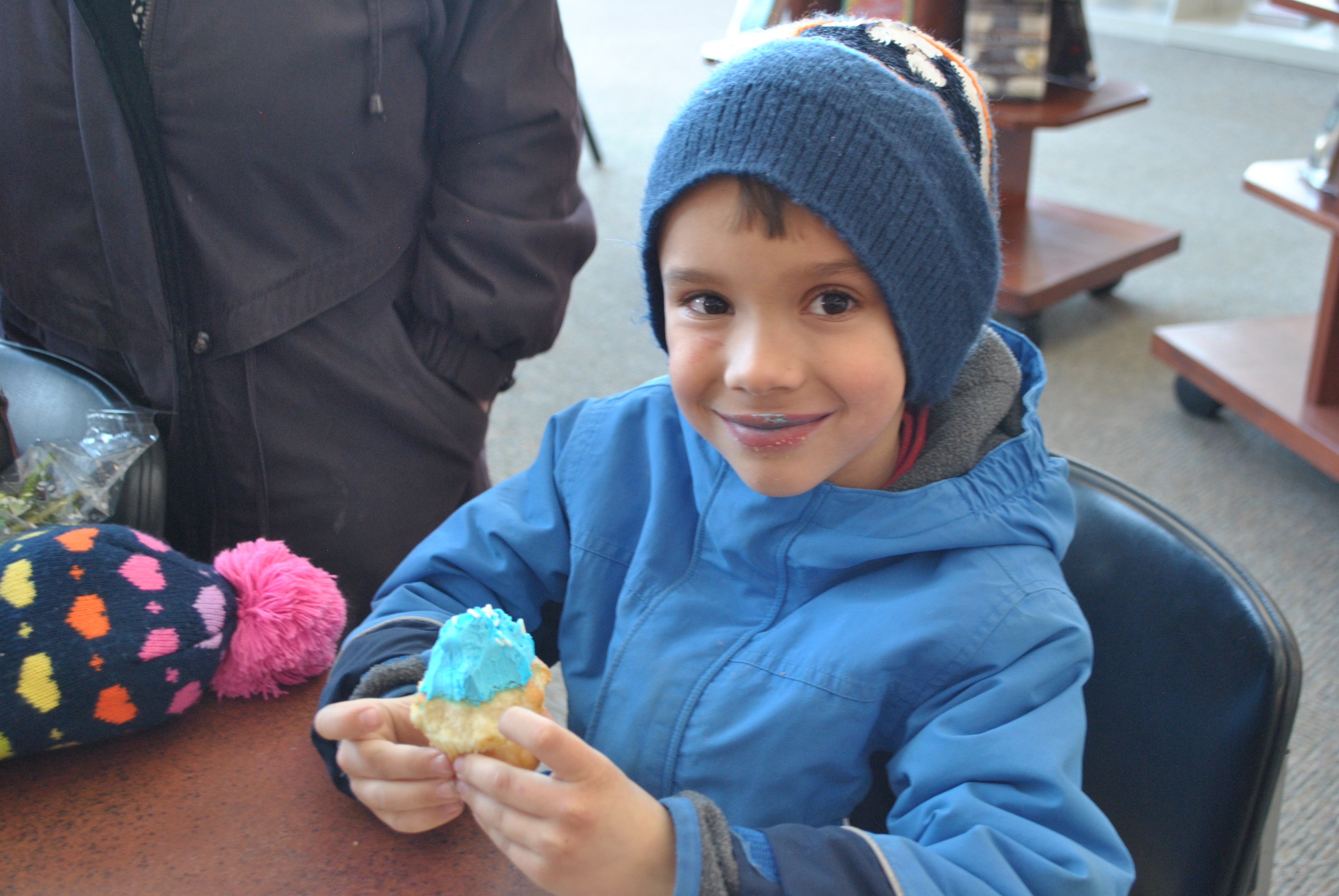 Child wearing blue coat and blue hat eats cupcake with blue icing