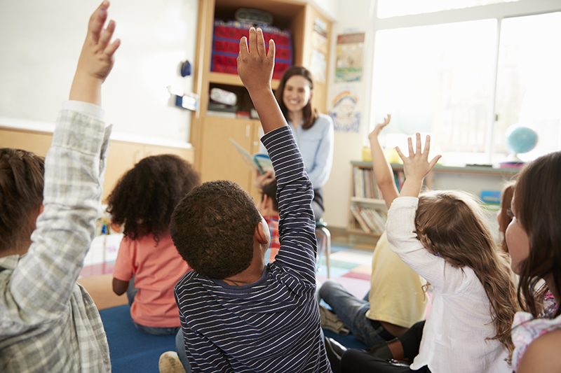Picture of a classroom showing children raising their hands and a smiling teacher at front of room